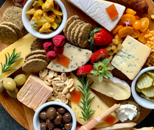 Assorted cheese and cracker platter with fruits, olives, and pickles on a wooden board.