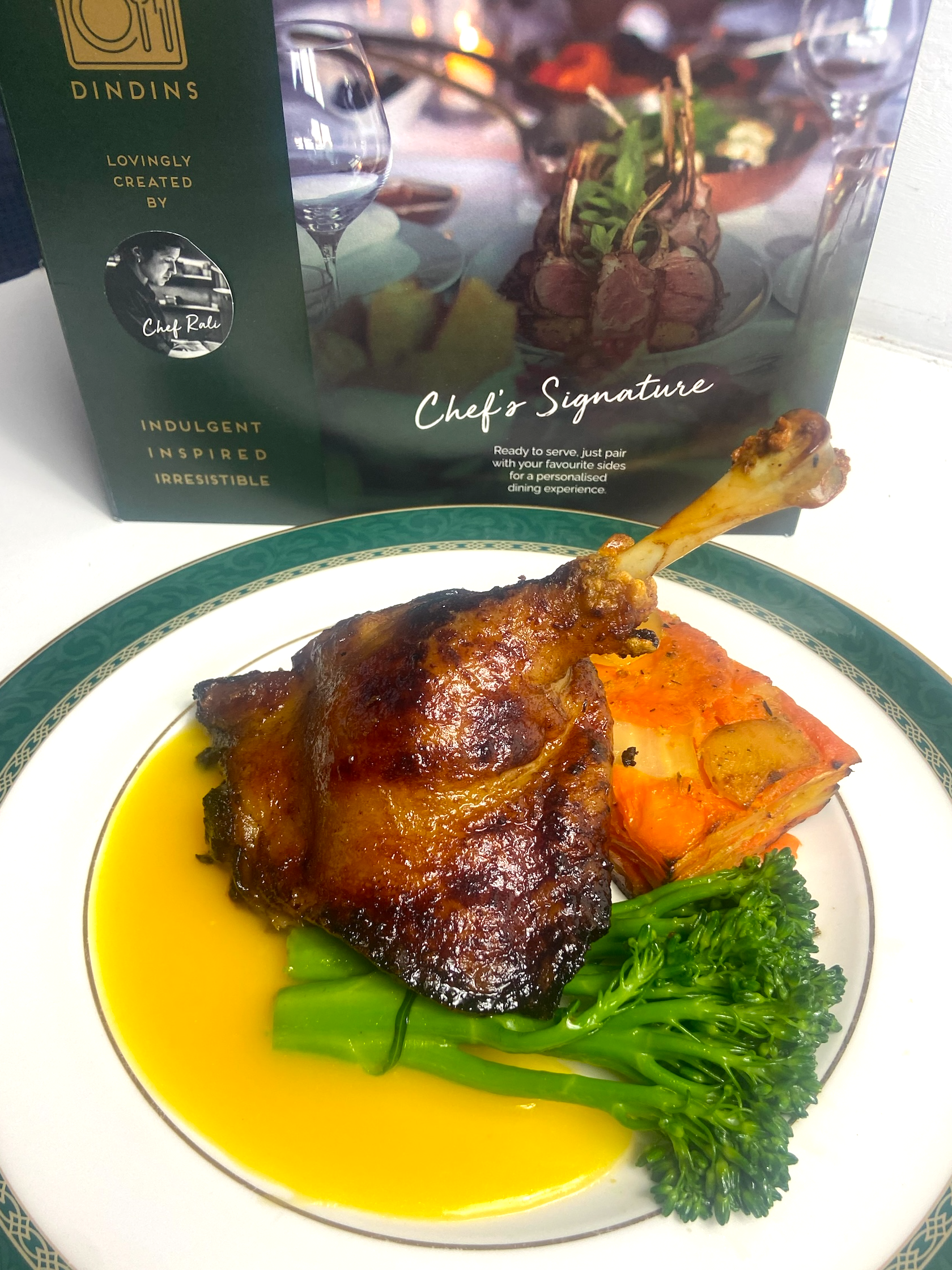 Plated dish with roasted leg of duck, vegetables, and sauce, with a branded box in the background.